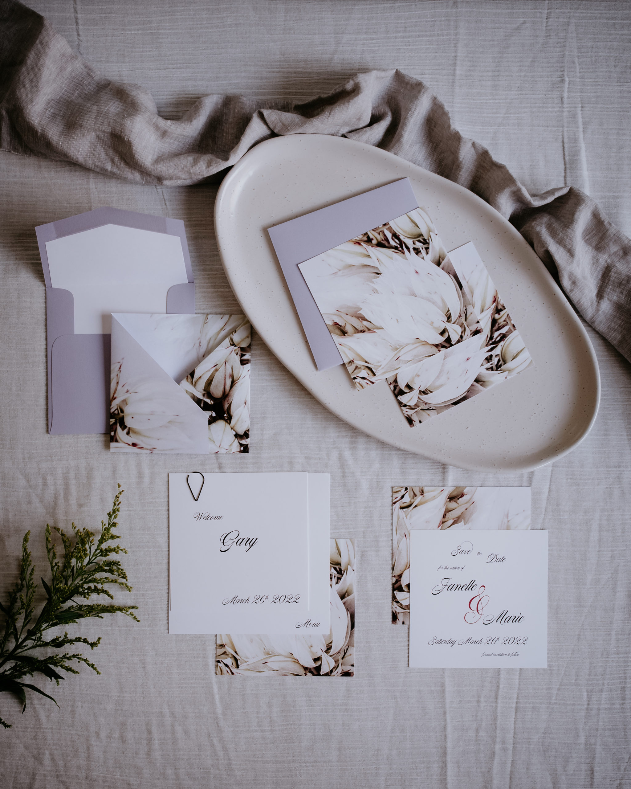 A delicate white and lilac floral wedding invitation with an origami fold. The invitation sits with the save the date card, and the menu and placecard.