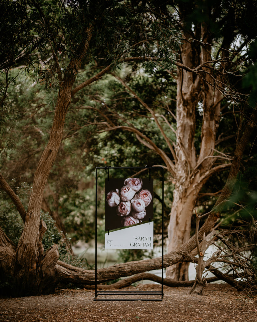 Large two-piece wedding welcome sign, with pink flowers and couples name. Sign is hung on a black stand and stands in front of a tree.