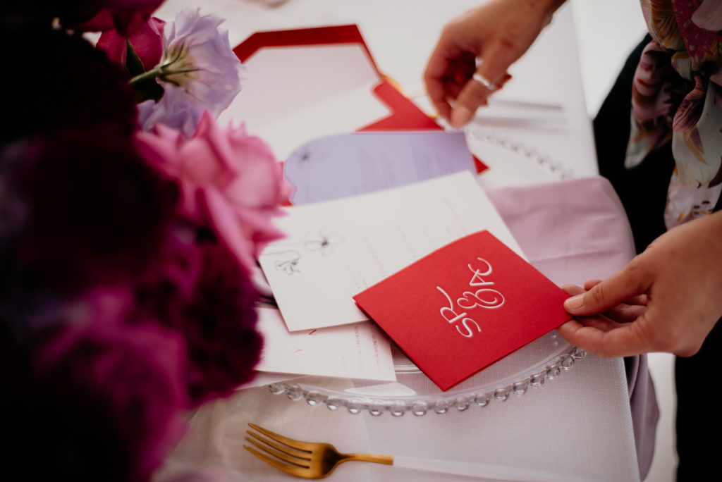 A bride holding a bold and colourful wedding invitation suite, with a red invitation pocket, a nude invitation and arched lavender wedding details card.