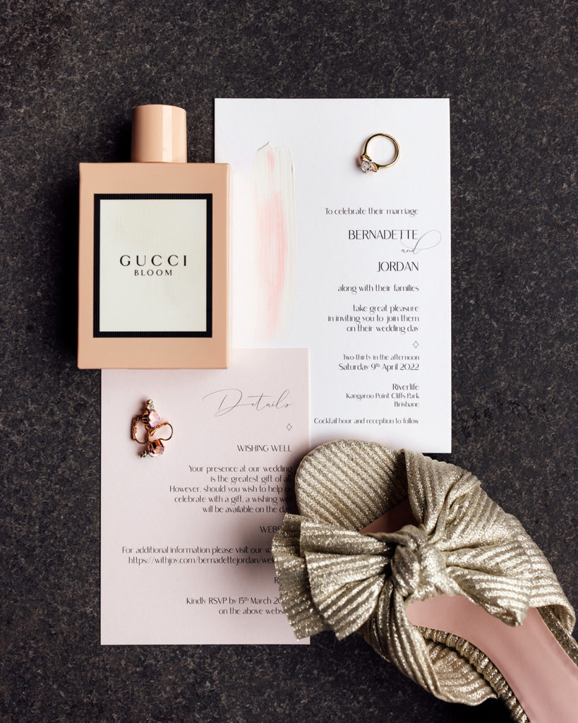 Pink and white delicate wedding invitations with wedding shoes, rings, and perfume.