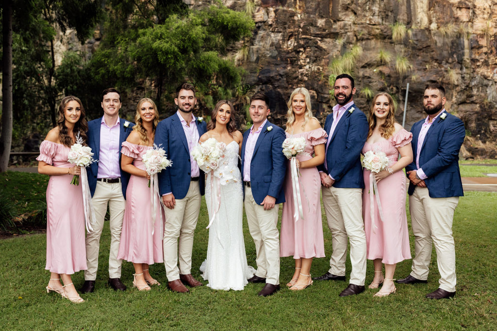 Bride and groom with bridal wedding party wearing pink bridesmaids dresses, and navy and brown suits.