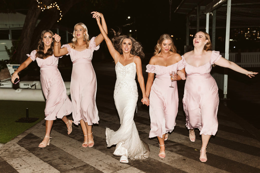 Bride in shoestring strap white dress with bridesmaids in pale pink dresses, walking along a pier at night holding hands.