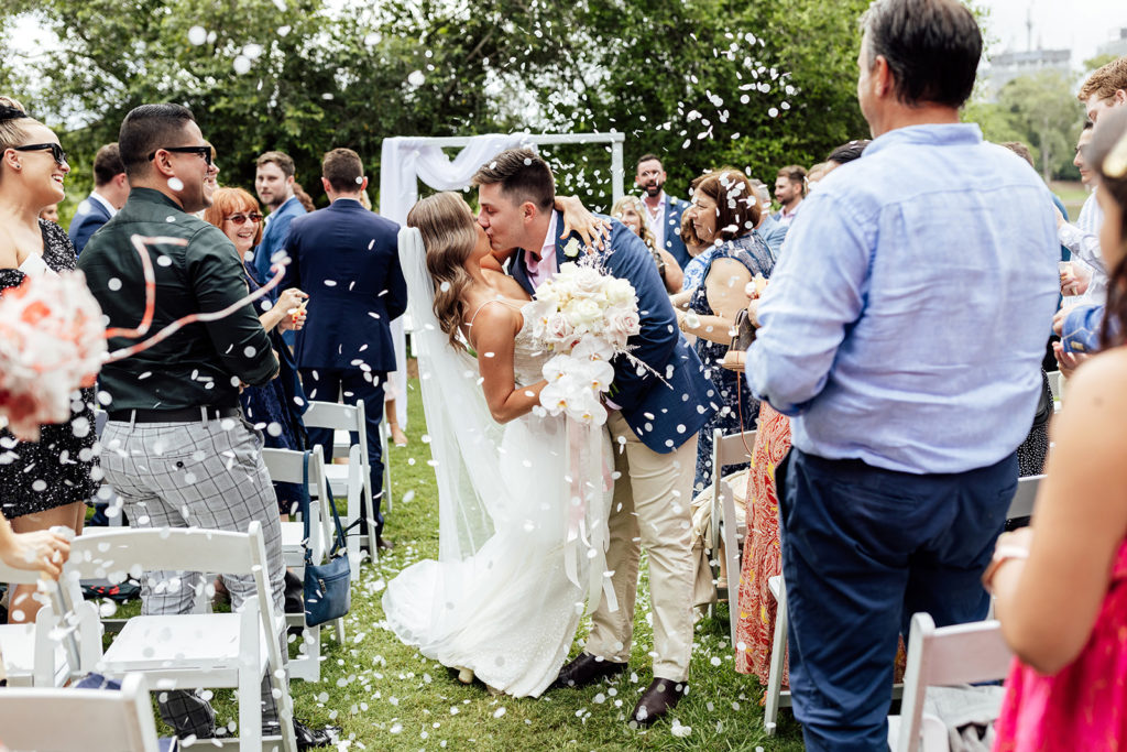 Bride and groom kissing, coming back down the aisle in a park with confetti and guests in the audience.