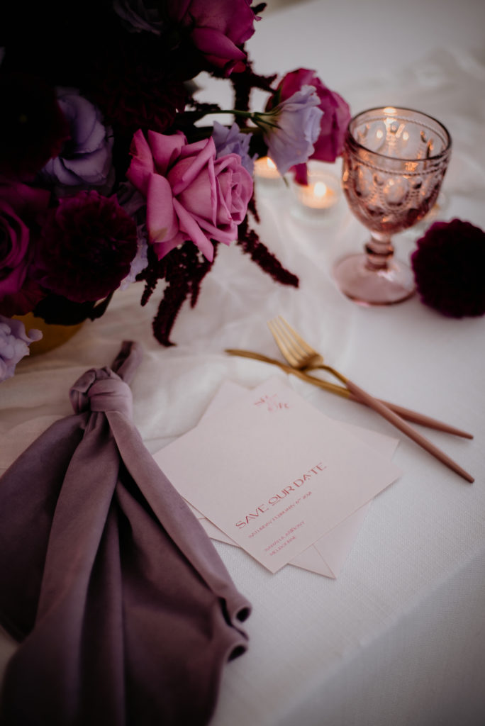 Save the date card with nude cardstock and red digital printing.  The save the date card lies on a table with a purple velvet napkin, gold cutlery, and pink and purple flowers.