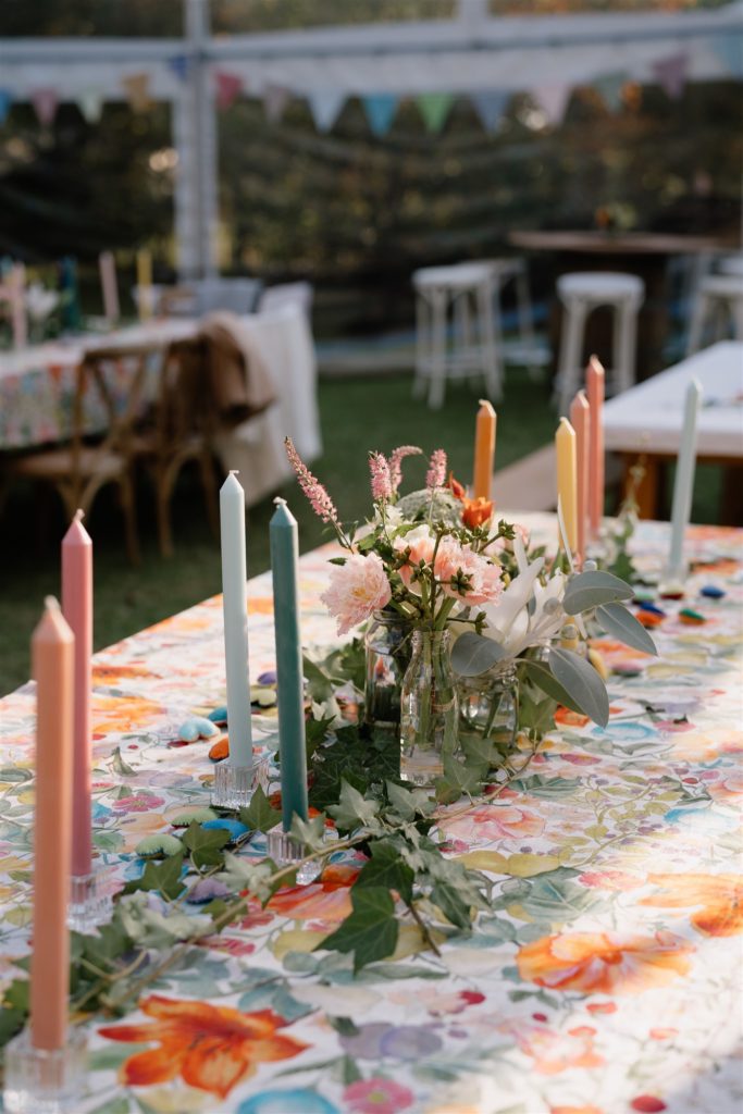 Wedding reception styling with orange and sage green candles and florals. A colourful floral tablecloth is on the table.