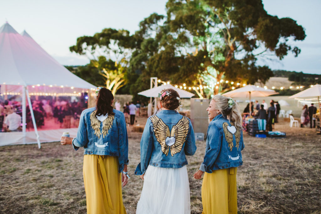 A bride and her two bridesmaids are standing wearing denim jackets at their festival style outdoor wedding. Wedding planner and stylist Paradise Hunter.