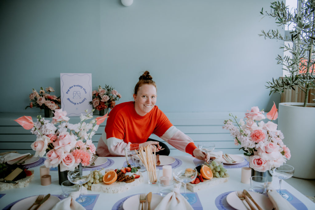 Wedding planner and stylist Caitlin from Paradise Hunter is pictured with a table that she styled for an event. The table is styled in bright pink and blues, with flowers, frit, and tableware.