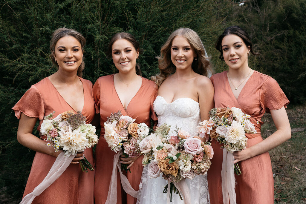 A bride wearing a white dress stands with her bridesmaids wearing terracotta orange dresses.  Bouquets by Melbourne wedding florist Oh Hello Petal.