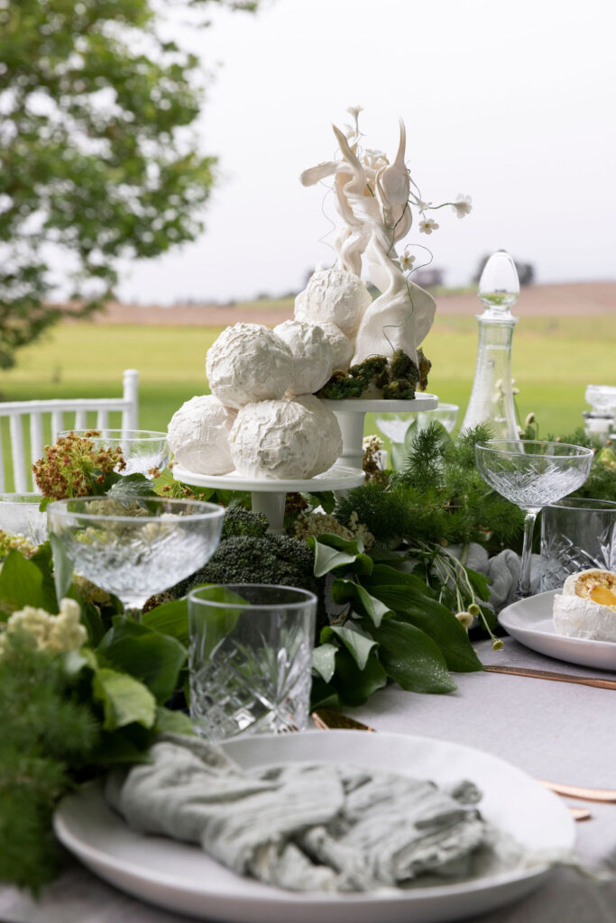 An artistic white wedding cake with natural shapes, sitting on a wedding reception table styled with greenery and clear glassware.