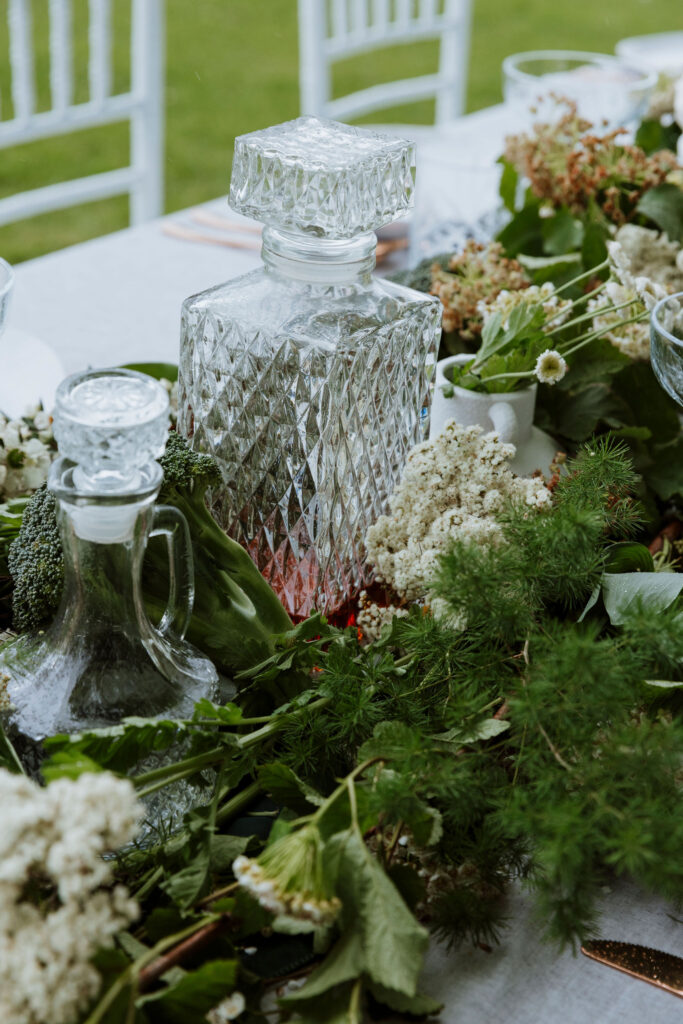 A crystal decanter sits on a wedding reception table styled with green foliage.
