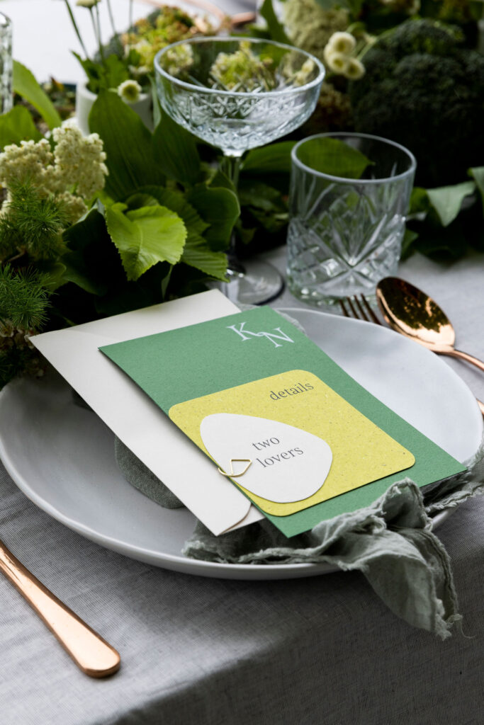Natural wedding invitations with green grass paper and a stone coloured card on the front, with a stone coloured envelope.  The invitations sit on a table styled with green foliage.