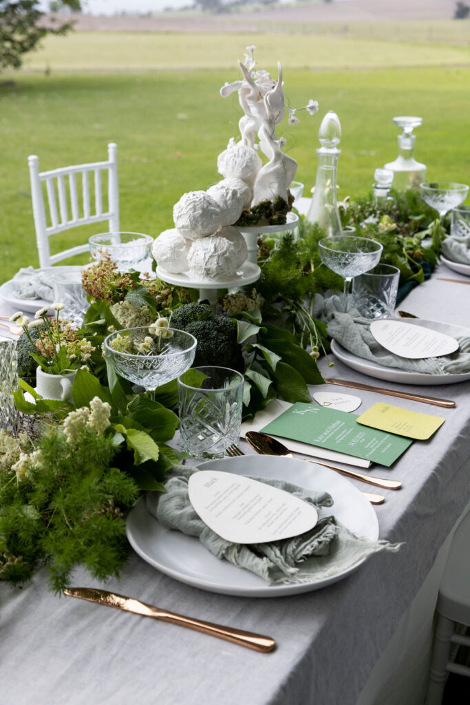 A green wedding reception with natural wedding invitations, green foliage, and a white wedding cake with natural shapes.