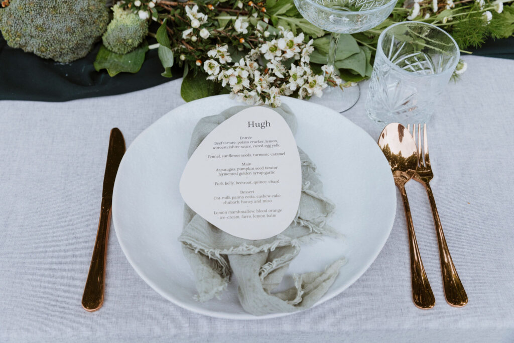 Pebble-shaped wedding menu with guest name on stone paper, sitting on a sage green linen napkin next to gold cutlery.