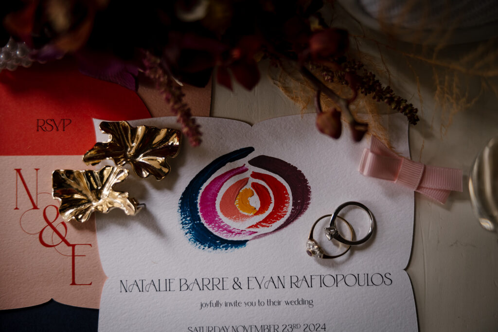 A flat lay of colourful painted wedding invitations with wedding rings and earrings.