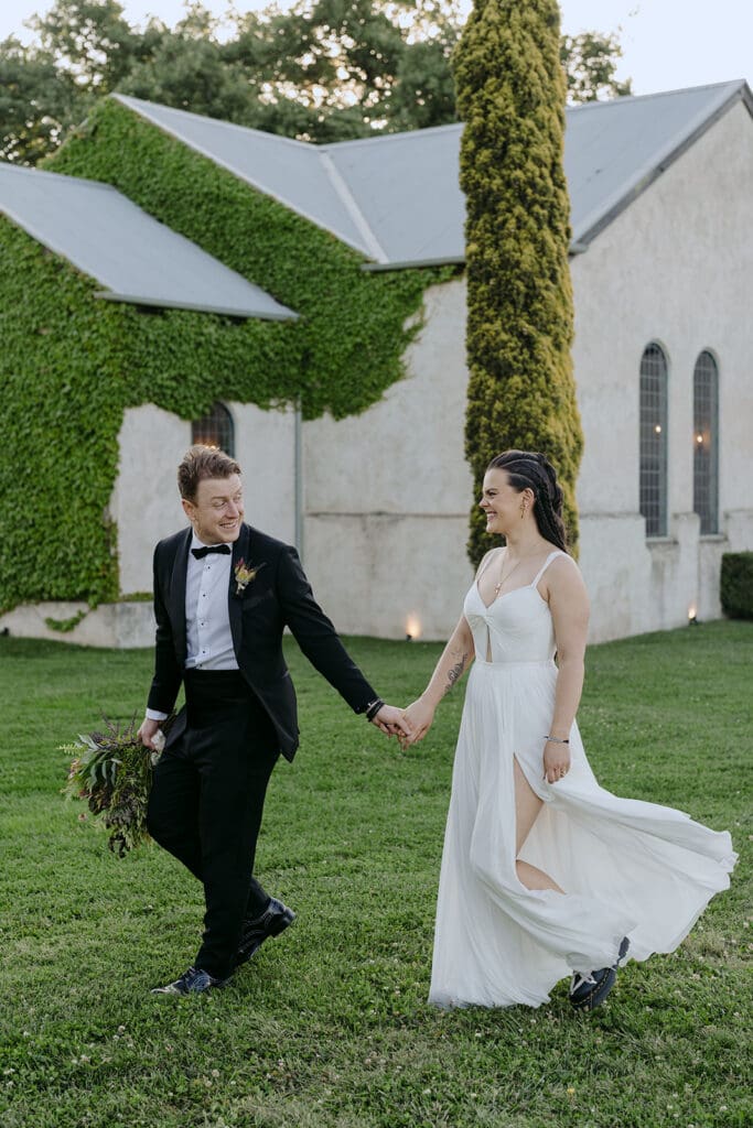 A bride and a groom holding hands in front of a chapel and walking along the grass