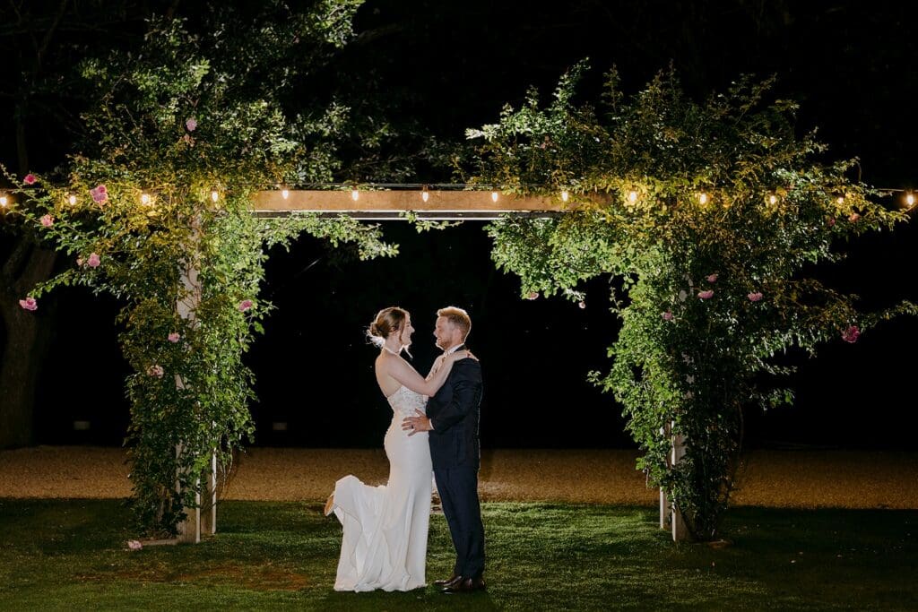 A bride and groom standing in front of an arbour, in the dark but backlit.