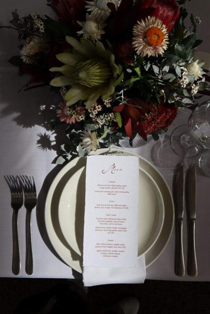 A wedding reception menu sitting on top of plates next to cutlery and native flowers.