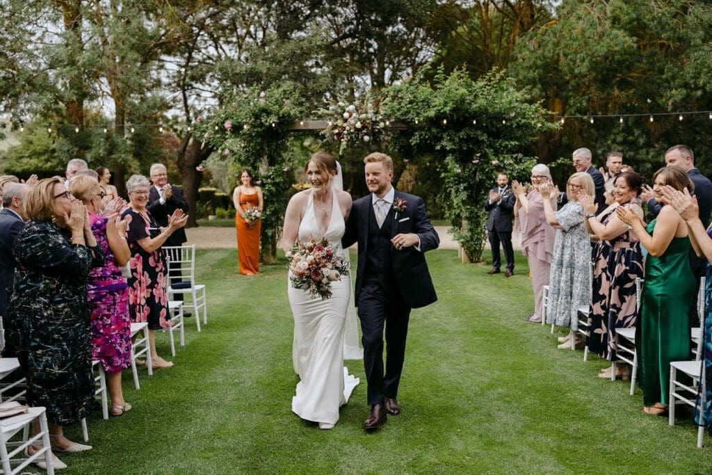 A couple walking back down the aisle after getting married on a lawn.