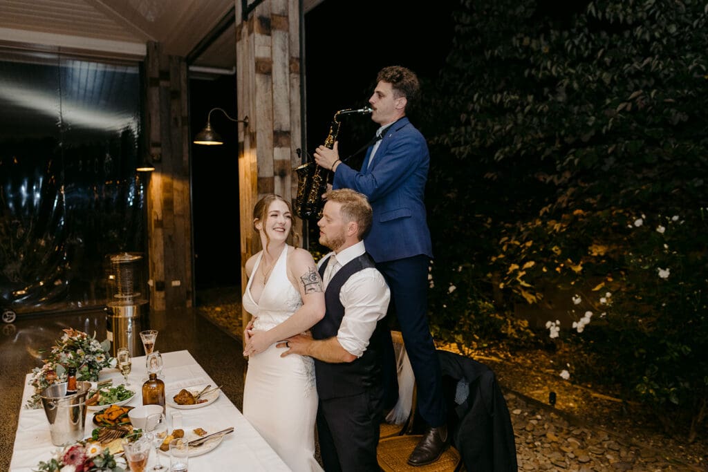 A bride and groom hugging at their wedding reception next to a roving saxophonist.